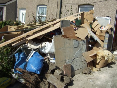 A large pile of mixed rubbish and waste materials situated on a pavement outside a residential building in Shoreditch. The foreground includes broken concrete blocks, wooden planks, and flattened packaging boxes made of cardboard. Some cardboard boxes are crushed and scattered, with a few partially collapsed or torn, showing brown and beige tones. There is also a blue plastic bag and white fabric or padding material among the debris. The waste extends outward, obstructing part of the sidewalk and spilling close to a wall of the building behind. The background features three residential windows with white frames, partially obscured by the rubbish, and a small shrub or plant visible at the left edge. The scene is outdoors during daylight, with natural sunlight casting shadows, and it appears to be an active rubbish disposal or collection area in Shoreditch, aligning with commercial waste removal services provided by Commercial Waste Shoreditch.