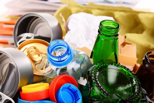 A close-up view of various types of recyclables and waste materials, including crushed aluminum cans in silver, a green glass bottle, a translucent plastic bottle with a blue cap, and colorful plastic bottle caps in red, yellow, and blue, all placed among crumpled white paper and brown cardboard packaging. The background shows a yellowish fabric or insulation material, suggesting an indoor or outdoor waste collection setting. The arrangement highlights the different textures and colors of recyclable containers, aligned with rubbish removal services in Shoreditch, showing the variety of waste that companies like Commercial Waste Shoreditch handle for efficient rubbish disposal and recycling efforts.