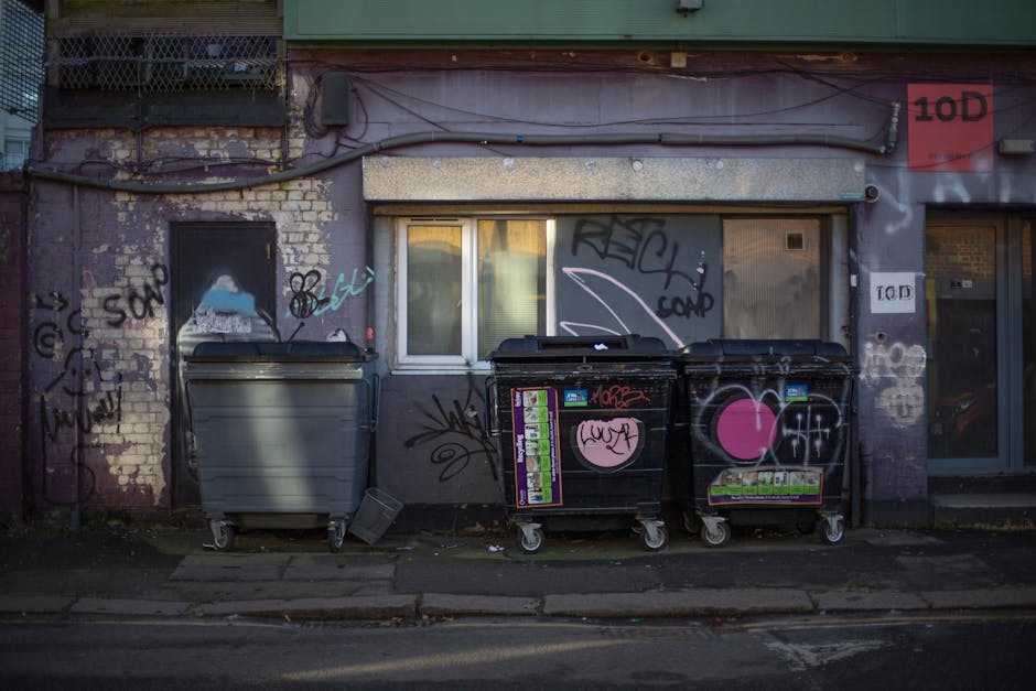 Three large waste containers are positioned along the sidewalk in front of a weathered, graffiti-covered brick building facade with a small, rectangular window and a door to the right. The first conta