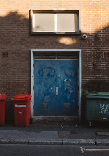 Three large waste containers are positioned along the sidewalk in front of a weathered, graffiti-covered brick building facade with a small, rectangular window and a door to the right. The first conta